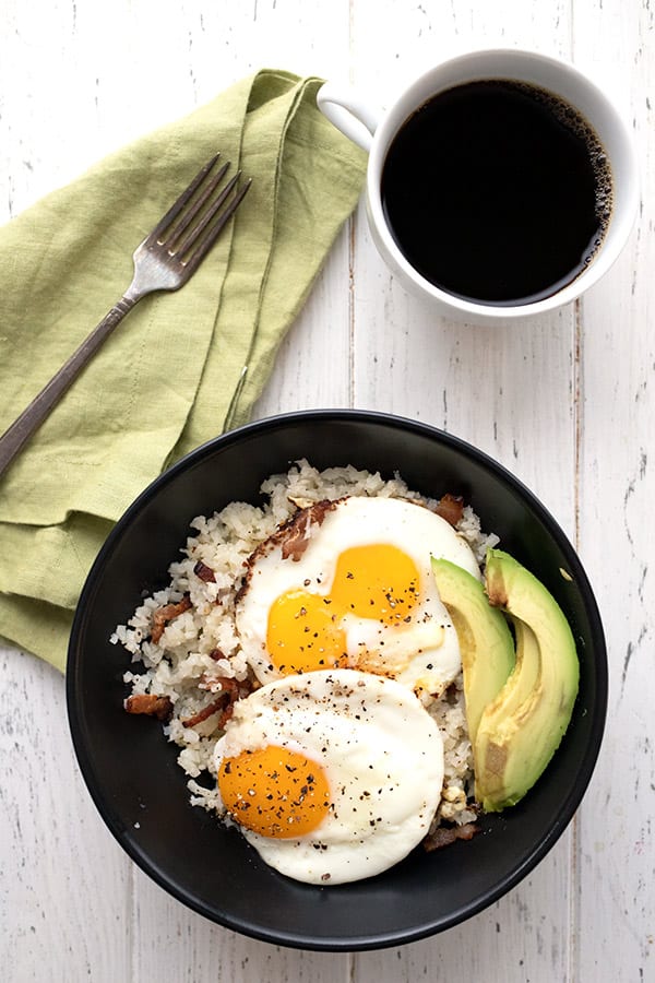 Top down photo of keto breakfast grits with bacon, fried eggs, and avocado in a black bowl. A cup of coffee and a green napkin with a fork are also featured.