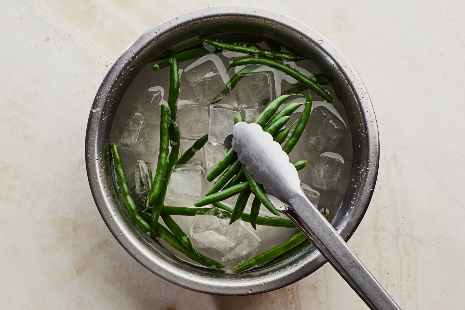 Shocking blanched green beans in a bowl of ice water using tongs.