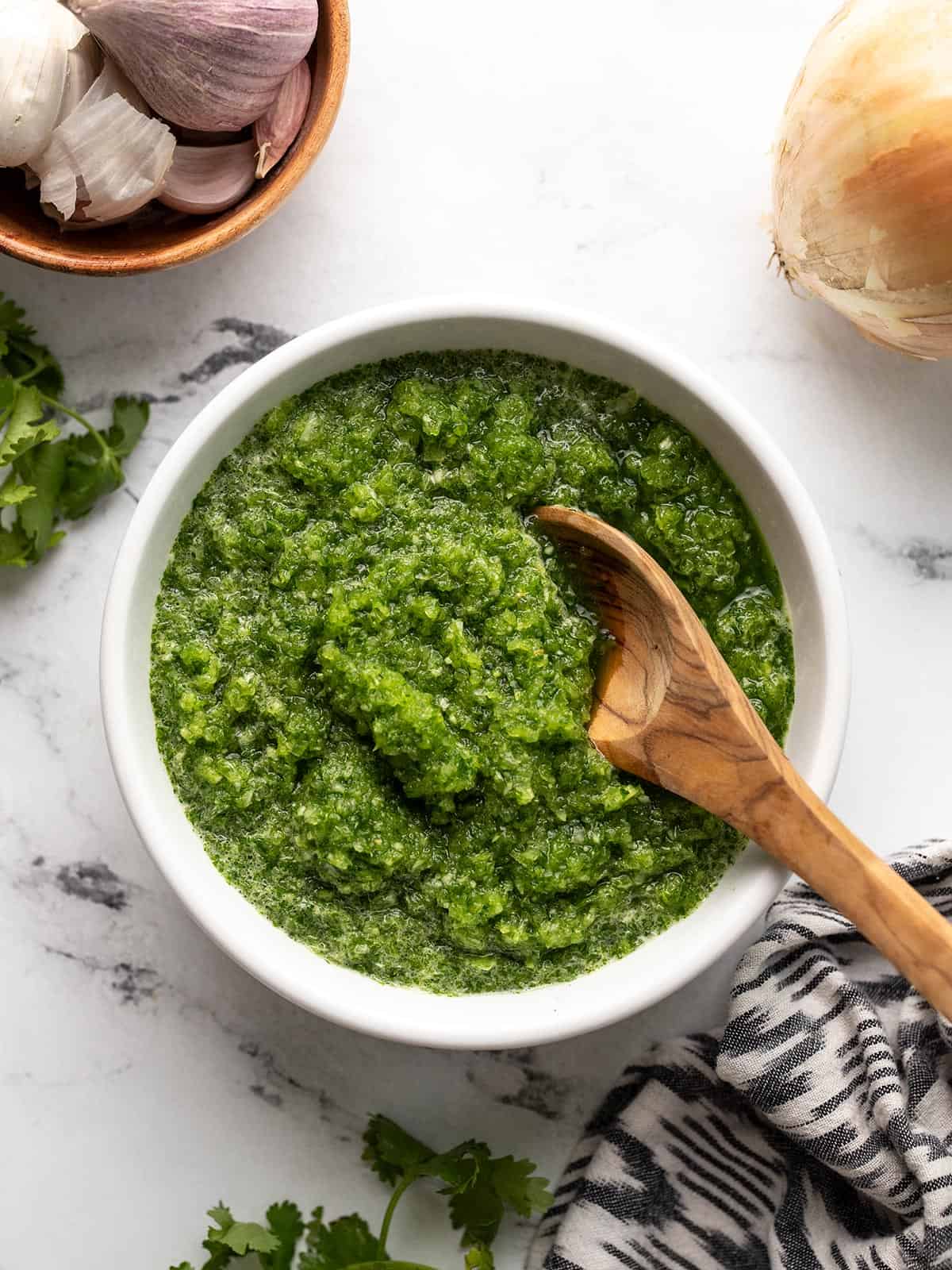 Overhead shot of green sofrito in a white bowl with a wooden spoon in it.