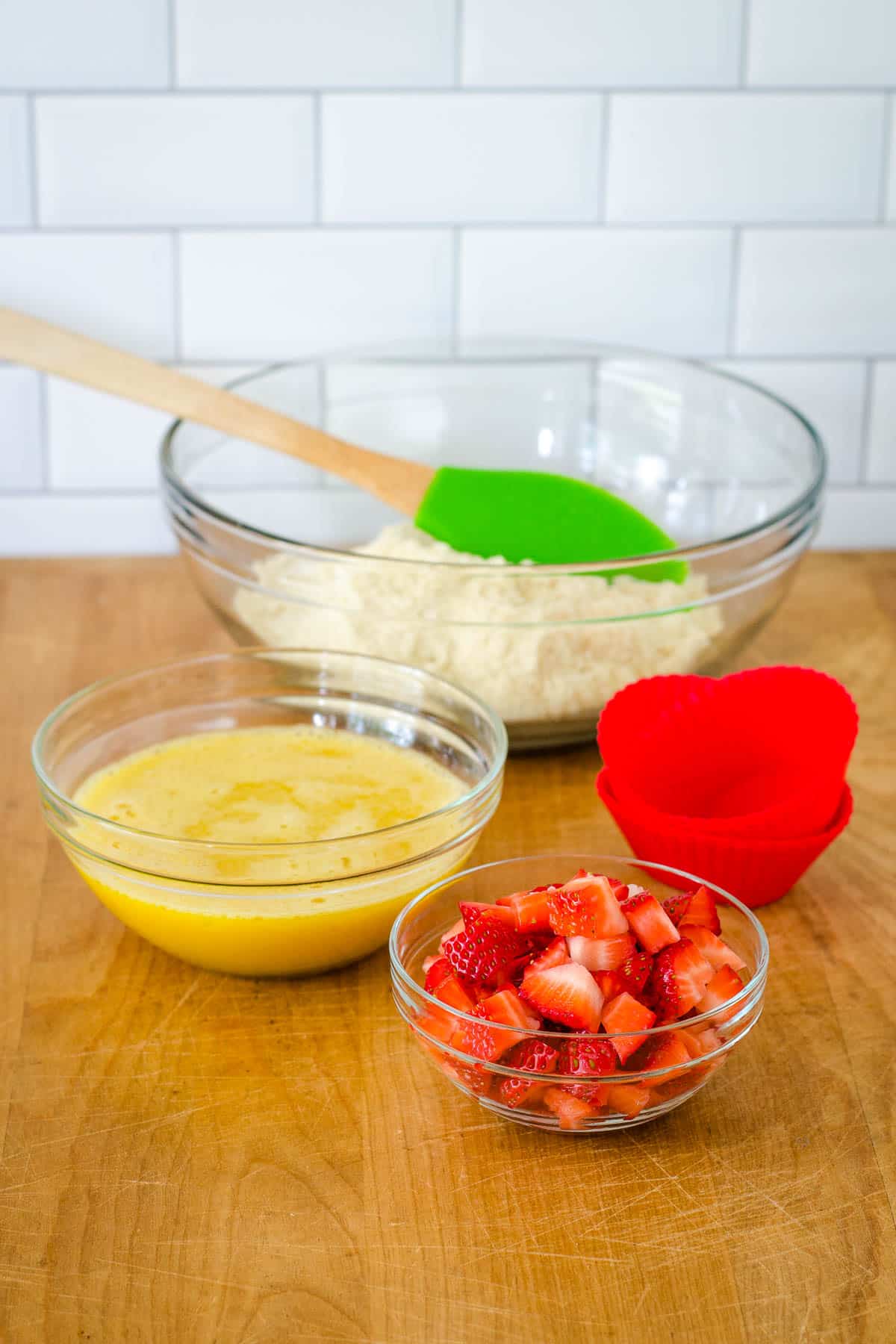 Bowls of dry ingredients, wet ingredients, chopped strawberries, red muffin cups