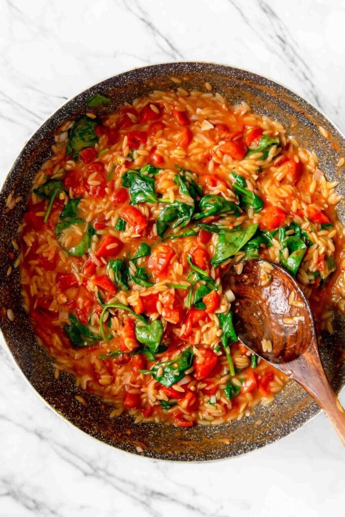 Large pan of tomato orzo with spinach with a wooden spoon resting in pan.