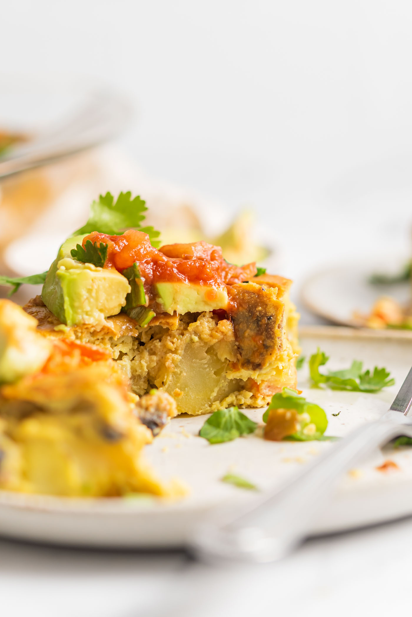 Slice of vegan eggless casserole with chunks of potato and veggies in it, topped with avocado and salsa.