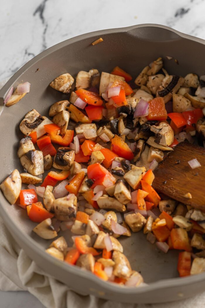 Mushroom, onion, and red bell pepper cooking in a skillet with a wooden spoon.