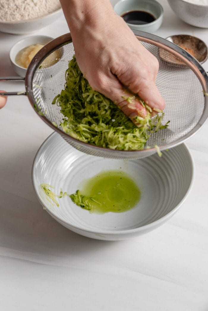 Hand squeezing grated zucchini over a strainer to remove excess water.