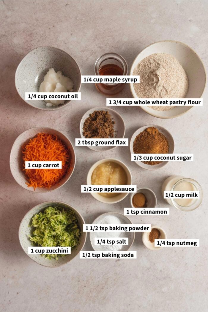 All the ingredients for making carrot zucchini muffins gathered in various small dishes. Each ingredient is labelled with text overlay.