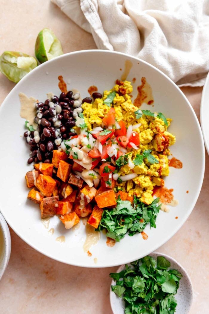 Breakfast burrito bowl with cilantro, tofu scramble, black beans, roasted sweet potato, chipotle sauce, and hot sauce drizzled over top.