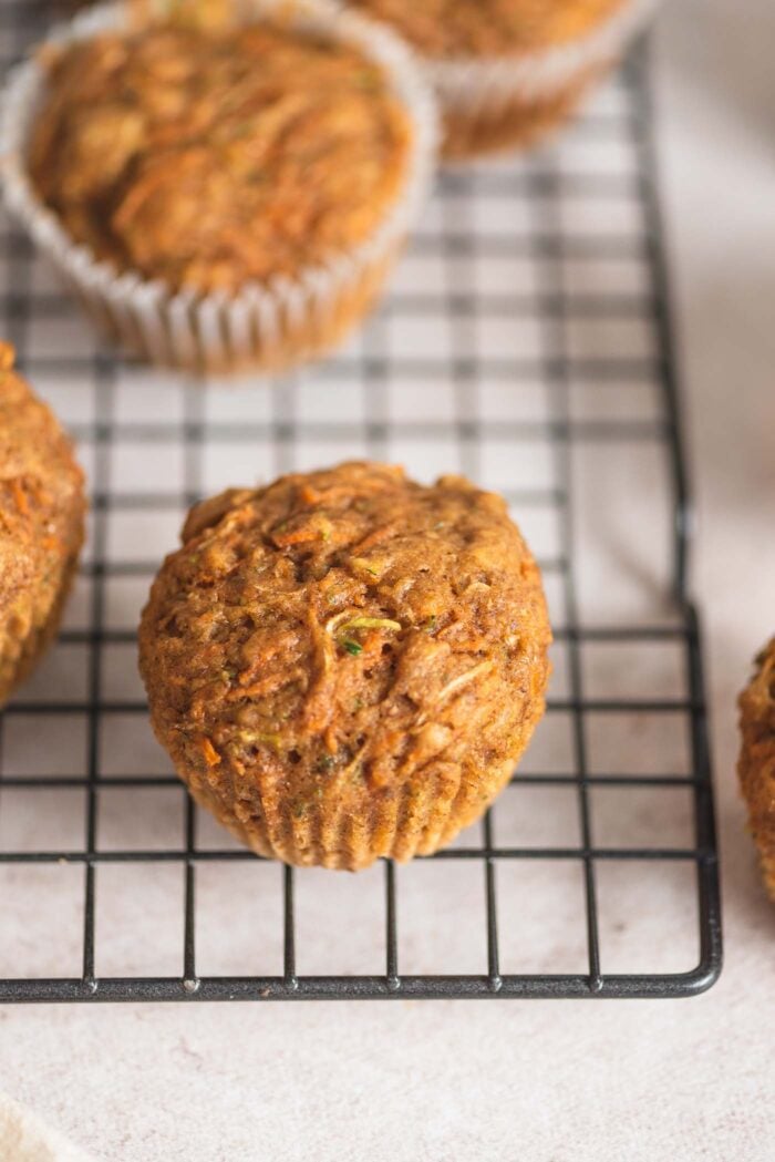 A small carrot zucchini muffin sitting on the edge of a baking rack with more muffins in the background.