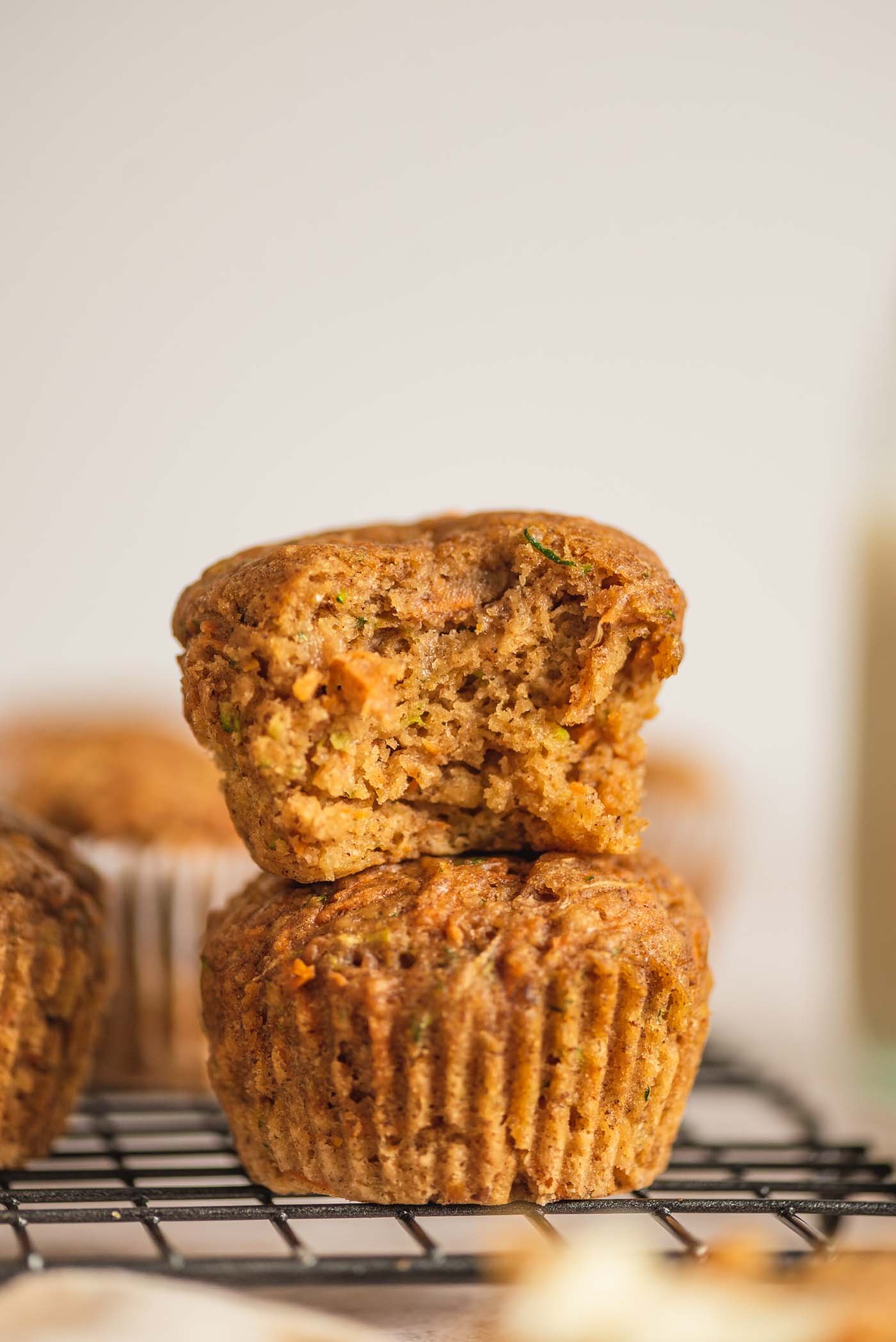 Two carrot zucchini muffins stacked on one another. The muffin on top has a bite out of it so you can see the texture and bits of grated carrot and zucchini inside.