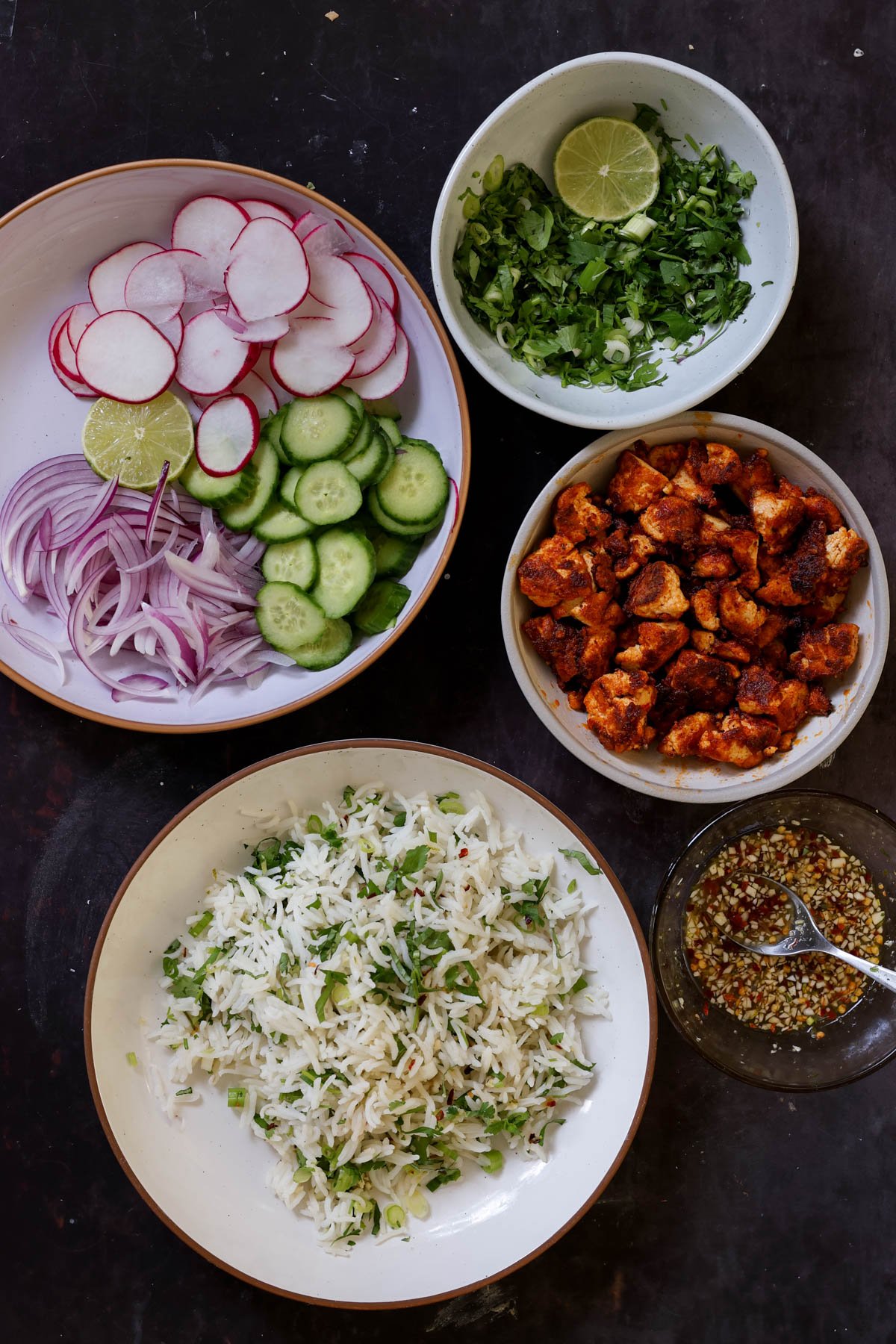 harissa tofu, herbed rice, fresh veggies, herbs, and dressing in bowls on a dark table