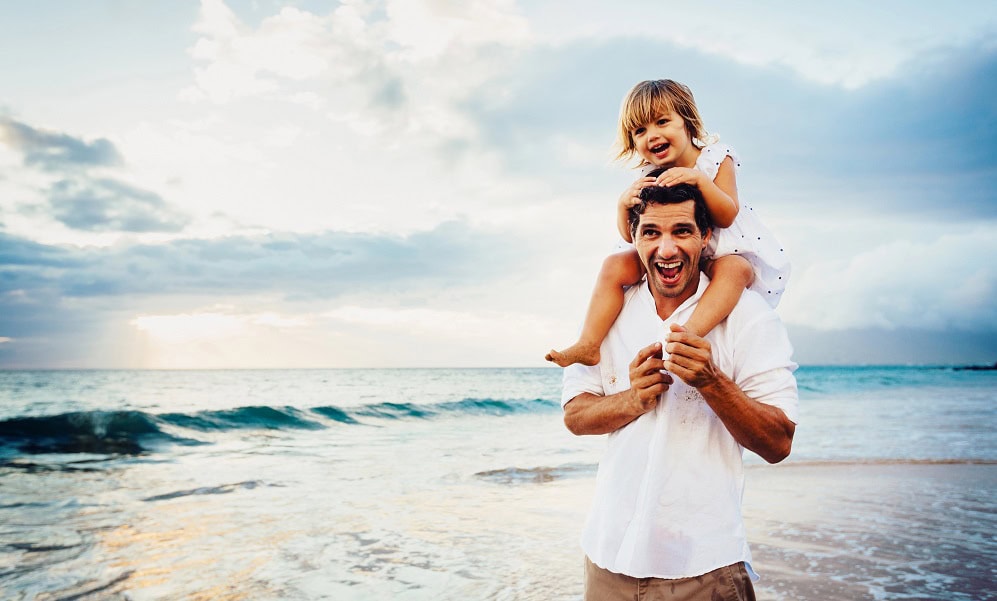 A happy dad with his daughter on his shoulders at the beach.